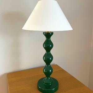 May include: A green table lamp with a white lampshade. The lamp has a round base and a curvy, stacked design. The lampshade is cone-shaped. The lamp is on a wooden table.