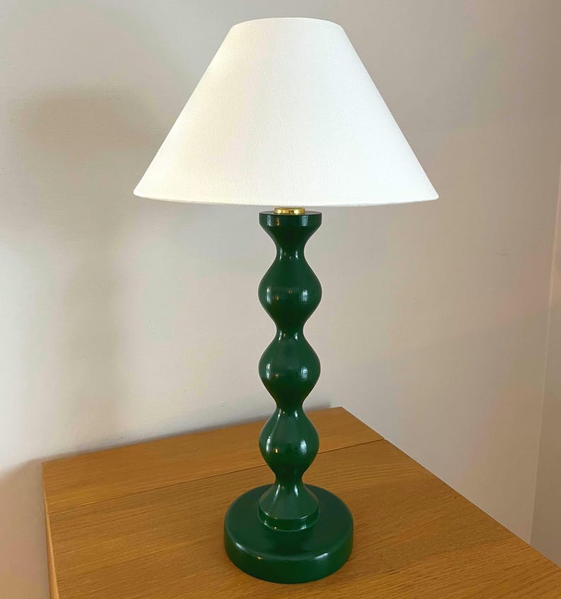 May include: A green table lamp with a white lampshade. The lamp has a round base and a curvy, stacked design. The lampshade is cone-shaped. The lamp is on a wooden table.