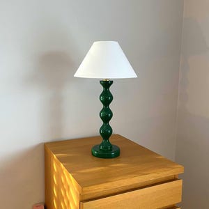 May include: A green table lamp with a white lampshade. The lamp features a curvy, stacked design and is placed on a wooden bedside table. The lamp's base is a deep green, and the lampshade is a simple, cone shape.