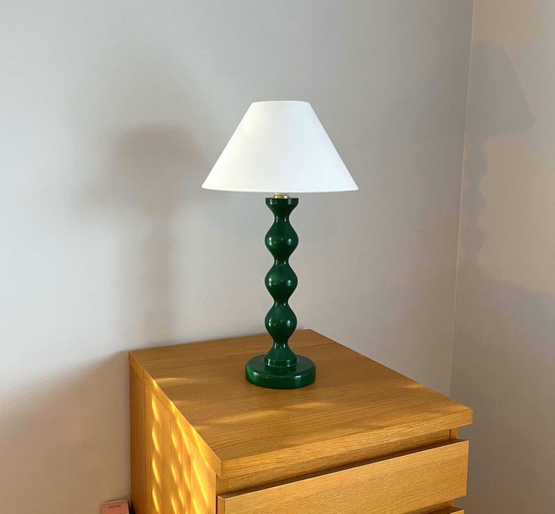 May include: A green table lamp with a white lampshade. The lamp has a curvy, stacked design and sits on a wooden bedside table. The lamp's base is a deep green colour, and the lampshade is a simple, cone shape.