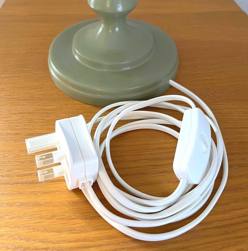 May include: A sage green lamp base with a white electrical cord, plug, and on/off switch. The lamp base is round with a decorative design. The cord is coiled on a wooden surface.