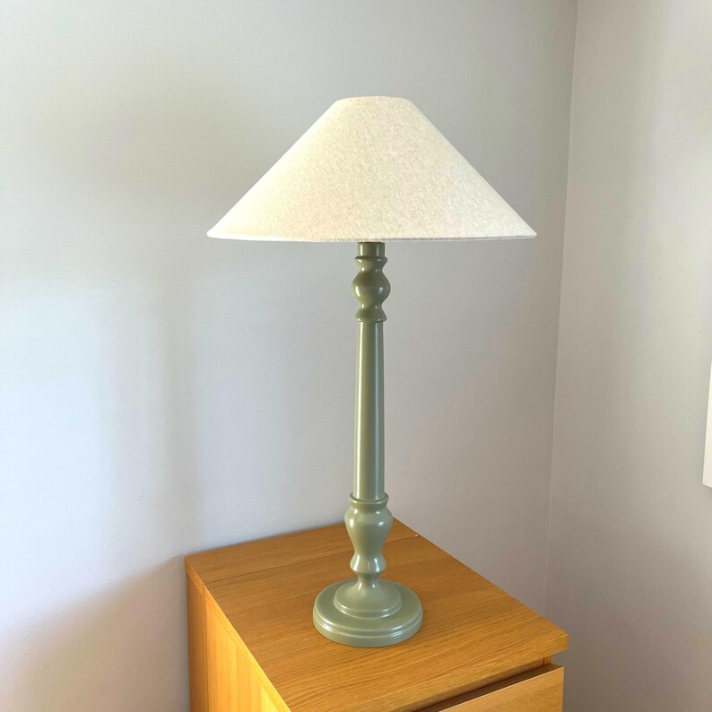 May include: A tall, sage green table lamp with a cream-colored lampshade. The lamp has a turned wooden base and a long, slender stem. The lampshade is conical in shape.
