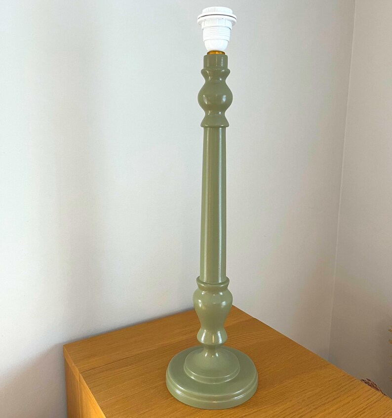 May include: A sage green table lamp base with a white bulb socket. The lamp has a round base, a long, slender stem, and decorative bulbous sections. The lamp is placed on a wooden surface.