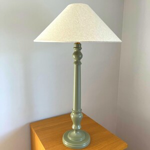 May include: A tall, sage green table lamp with a white lampshade. The lamp has a turned wooden base and a long, slender stem. The lampshade is a light, neutral color and is triangular in shape. The lamp is sitting on a wooden surface.