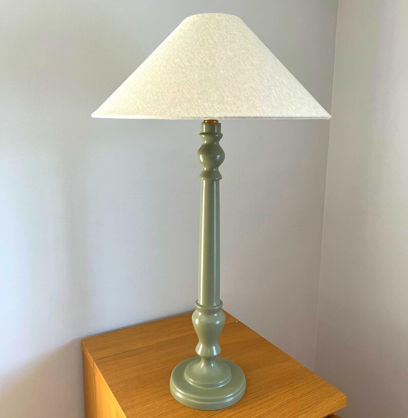 May include: A tall, sage green table lamp with a white lampshade. The lamp has a turned wooden base and a long, slender stem. The lampshade is a light, neutral color and is triangular in shape. The lamp is sitting on a wooden surface.