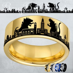 May include: A gold-colored ring with a black cityscape design featuring Godzilla silhouettes. The ring has a polished finish and a wide band. Additional rings in black, silver, and blue are shown below the main ring.