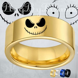 May include: A gold-colored ring with a black Jack Skellington face design. The ring has a flat, wide band. Additional rings in gold, silver, blue, and black are shown at the bottom. The background features a stylized face design.