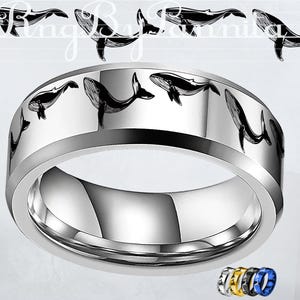 May include: A silver-colored ring with a polished finish. The ring features a repeating pattern of black whale illustrations around the band. The ring has a wide band design and a smooth, reflective surface. Additional rings in gold, black, and blue are shown.