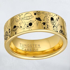 May include: A gold-colored tungsten carbide ring with a detailed space-themed design. The ring features black line art illustrations of planets, UFOs, and other space elements. The text "Tungsten Carbide" is engraved on the ring.