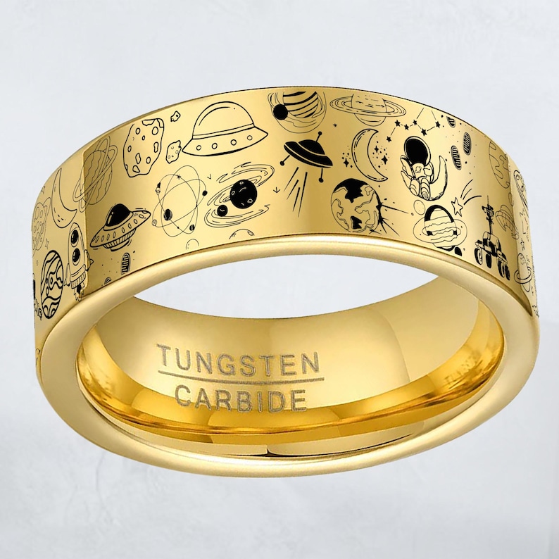 May include: A gold-colored tungsten carbide ring with a detailed space-themed design. The ring features black line art illustrations of planets, UFOs, and other space elements. The text "Tungsten Carbide" is engraved on the ring.