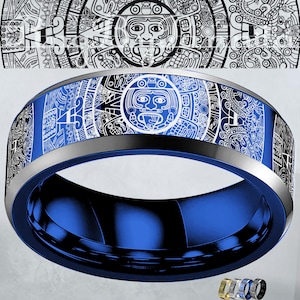 Op de afbeelding: Een blauwe ring met een gedetailleerd wit en zwart Azteeks kalenderontwerp. De ring heeft een gepolijste blauwe binnenkant en een zilverkleurige rand. Het ontwerp toont een centraal gezicht en ingewikkelde patronen.