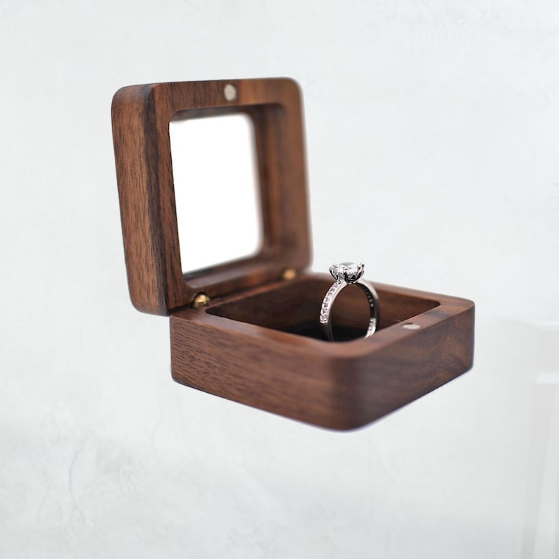 May include: A square, open wooden ring box with a clear top, displaying a diamond ring. The box is made of dark brown wood and has a hinged lid. The ring features a large, round-cut diamond.