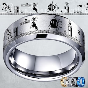 Puede incluir: Un anillo plateado con un acabado de metal cepillado. El anillo presenta un diseño grabado en blanco y negro de una escena de videojuego con un personaje, setas y una bomba. El anillo tiene un interior pulido y una selección de anillos de colores en la parte inferior.