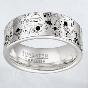May include: A silver tungsten carbide ring with a detailed black and white space-themed design. The ring features illustrations of planets, rockets, UFOs, and astronauts. The words "TUNGSTEN CARBIDE" are engraved on the inside of the ring.