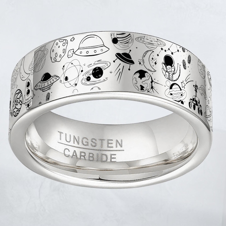 May include: A silver tungsten carbide ring with a detailed black and white space-themed design. The ring features illustrations of planets, rockets, UFOs, and astronauts. The words "TUNGSTEN CARBIDE" are engraved on the inside of the ring.