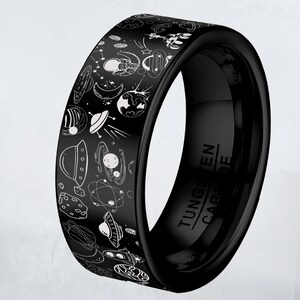 May include: Black tungsten carbide ring with a space-themed design. The ring features white illustrations of planets, stars, astronauts, and UFOs. The words "Tungsten Carbide" are engraved on the inside of the ring.