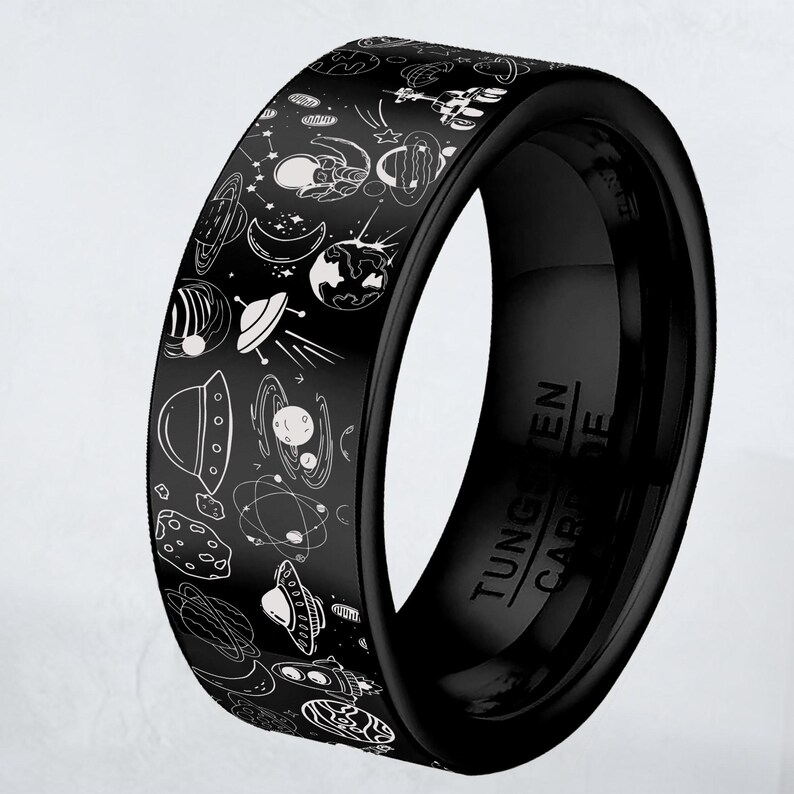 May include: Black tungsten carbide ring with a space-themed design. The ring features white illustrations of planets, stars, astronauts, and UFOs. The words "Tungsten Carbide" are engraved on the inside of the ring.