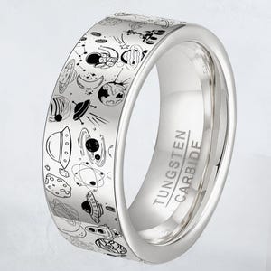May include: A silver tungsten carbide ring with a detailed outer band featuring a black and white space-themed design. The design includes planets, rockets, astronauts, and other celestial elements. The ring has the words "Tungsten Carbide" engraved on the side.