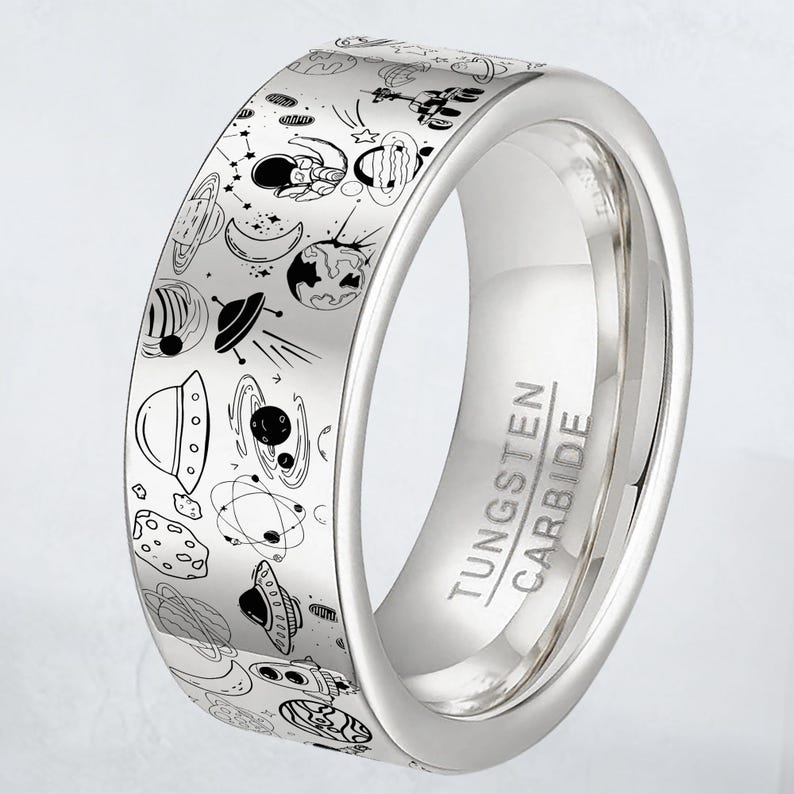 May include: A silver tungsten carbide ring with a detailed outer band featuring a black and white space-themed design. The design includes planets, rockets, astronauts, and other celestial elements. The ring has the words "Tungsten Carbide" engraved on the side.