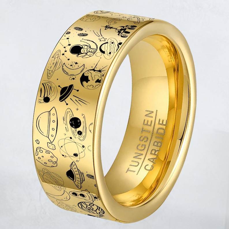 May include: A gold-colored tungsten carbide ring with a black space-themed design. The ring features illustrations of planets, astronauts, and UFOs. The text "TUNGSTEN CARBIDE" is engraved on the side of the ring.
