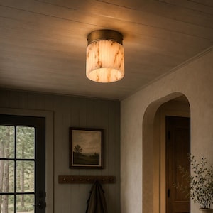 May include: A flush mount ceiling light fixture featuring a cylindrical shade made of translucent, cream-colored stone with brown veining. The shade is capped with a circular, metallic bronze-toned base that attaches directly to the ceiling.
