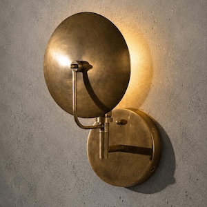 Puede incluir: Un aplique de pared fabricado en latón cepillado, con una placa de montaje circular y una pantalla ajustable en forma de disco. La lámpara emite un brillo cálido y suave desde detrás de la pantalla, creando un efecto de halo en la pared.