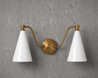 May include: A double wall sconce featuring a central circular brass mounting plate with two curved brass arms extending outward. Each arm supports a white conical shade pointing downward, creating a symmetrical lighting fixture design.