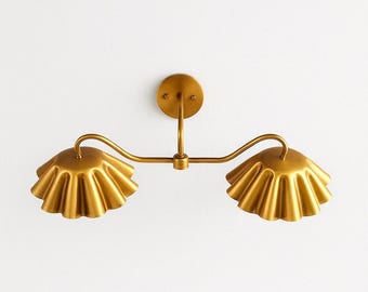 May include: A gold-colored wall sconce with two ruffled, bell-shaped shades. The fixture has a curved arm design and a circular base for mounting. The sconce is made of metal and has a warm, antique finish, suitable for interior lighting.