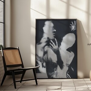 May include: Black and white framed art print featuring an abstract image of two figures. The artwork has a blurred, ethereal quality, with visible hands and faces. The print is displayed in a room with a black chair and a window.