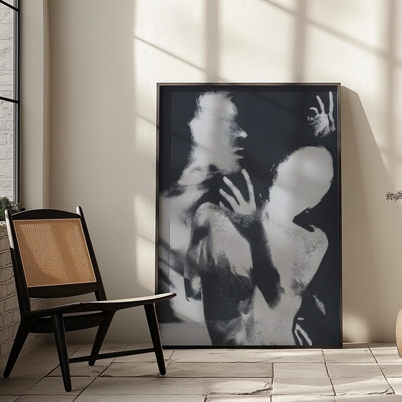 May include: Black and white framed art print featuring an abstract image of two figures. The artwork has a blurred, ethereal quality, with visible hands and faces. The print is displayed in a room with a black chair and a window.