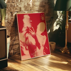 May include: A framed art print featuring a red and peach-toned image of two figures in an embrace. The artwork is displayed on a wooden floor, leaning against a brick wall, with a gold frame.