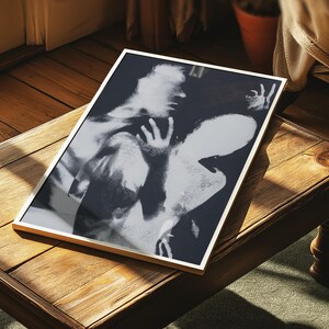 May include: Black and white framed art print featuring an abstract image of two figures. The artwork is displayed on a wooden table, with a gold-colored frame. The print has a high contrast, with areas of light and shadow.