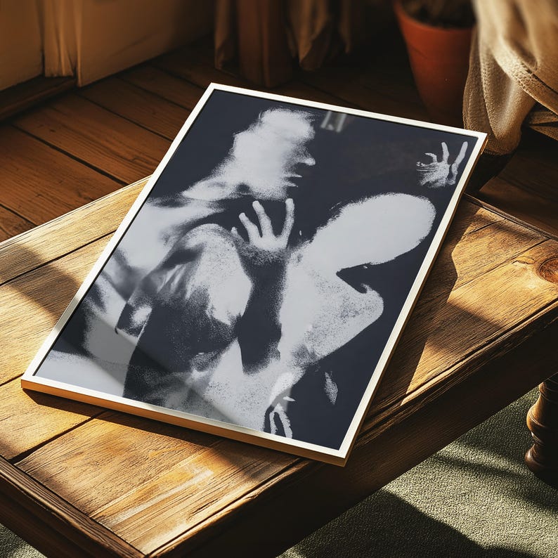 May include: Black and white framed art print featuring an abstract image of two figures. The artwork is displayed on a wooden table, with a gold-colored frame. The print has a high contrast, with areas of light and shadow.