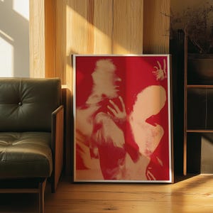 May include: A framed art print featuring a red background with two figures in a soft, beige hue. The figures are embracing, with one hand raised. The artwork is displayed on a wooden floor, next to a green leather couch.