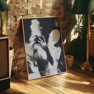 May include: A framed black and white art print depicting two figures in an embrace. The artwork is in a gold frame and leans against a brick wall and wooden floor. A green lamp and potted plant are in the background.