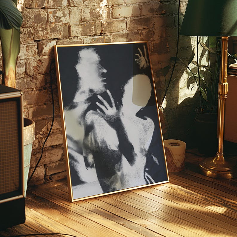 May include: A framed black and white art print depicting two figures in an embrace. The artwork is in a gold frame and leans against a brick wall and wooden floor. A green lamp and potted plant are in the background.