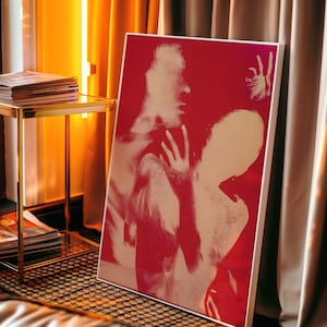 May include: A framed art print with a red background and a white and beige abstract image of two figures. The artwork is leaning against a wall near a gold side table and a window with yellow curtains.