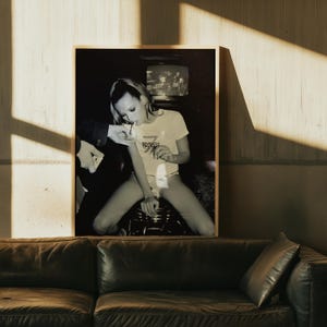 May include: Black and white framed print showing a person in a white t-shirt with the text "SEVENTH" and a person in a suit. The print is displayed above a dark brown leather sofa.