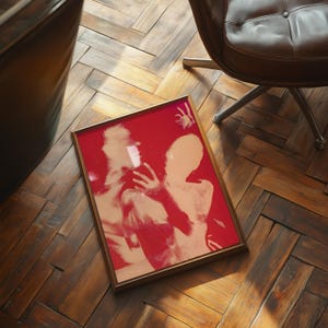 May include: A framed artwork featuring a red background with two nude figures in a soft, peach hue. The artwork is in a gold-colored frame and rests on a wooden floor. The image is a pop art style.