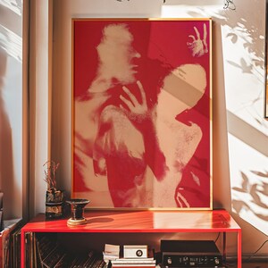 May include: A framed art print featuring a red and beige abstract design. The artwork depicts two figures in a close embrace, with a bold, graphic style. The print is displayed on a red cabinet, adding a pop of color to the room.