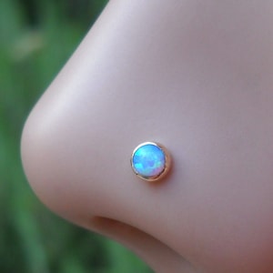 May include: A close-up of a nose piercing with a small, round, iridescent opal gemstone set in a gold setting.