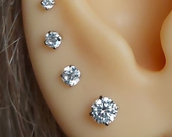 May include: Four clear, round-cut stud earrings of varying sizes are displayed on an ear. The earrings are set in silver-colored metal. The ear is light-toned, with visible curves and folds.
