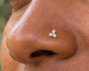 May include: A gold nose stud featuring three small, clear, round-cut stones arranged in a triangular cluster. The jewelry is set in a metallic gold base and is positioned on the side of a nostril, showcasing a simple and reflective design.