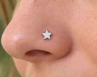 May include: A small, polished silver-toned star-shaped nose stud resting on the side of a nostril. The jewelry features a flat, five-pointed star design with a smooth, reflective metallic surface, securely positioned on the skin.