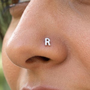 May include: A small, silver-toned metal nose stud featuring the capital letter R in a clean, sans-serif font. The piece is positioned on the side of a nostril, showcasing a polished, reflective surface and a simple, minimalist design.