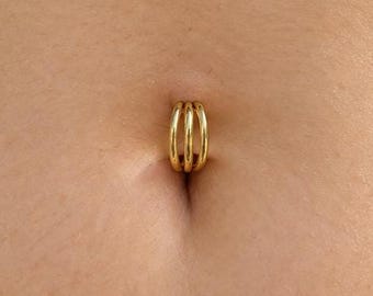 May include: A gold-toned triple-hoop belly button ring featuring three smooth, interconnected circular bands. The jewelry is positioned within the navel, showcasing a polished metallic finish and a simple, layered design structure.