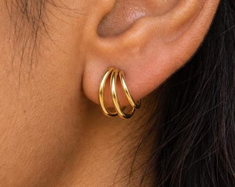 May include: A gold-toned triple hoop earring featuring three thin, polished circular bands that are connected at the base, creating a layered appearance when worn in the earlobe. The metal has a smooth, reflective finish.