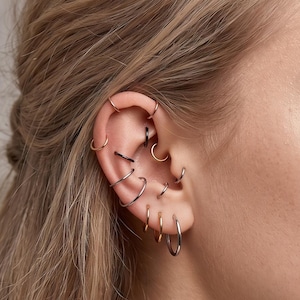 May include: A collection of multiple small hoop earrings worn in various piercings along the ear cartilage and lobe. The set includes thin metallic rings in silver, gold, and black tones, arranged in a layered stack across the ear structure.