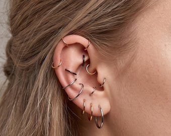 May include: A collection of multiple small hoop earrings worn in various piercings along the ear cartilage and lobe. The set includes thin metallic rings in silver, gold, and black tones, arranged in a layered stack across the ear structure.