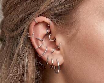 May include: A collection of multiple silver hoop earrings worn along the ear cartilage and lobe. The set includes various sizes of thin, circular metal rings positioned in several piercings, creating a layered look across the entire ear structure.